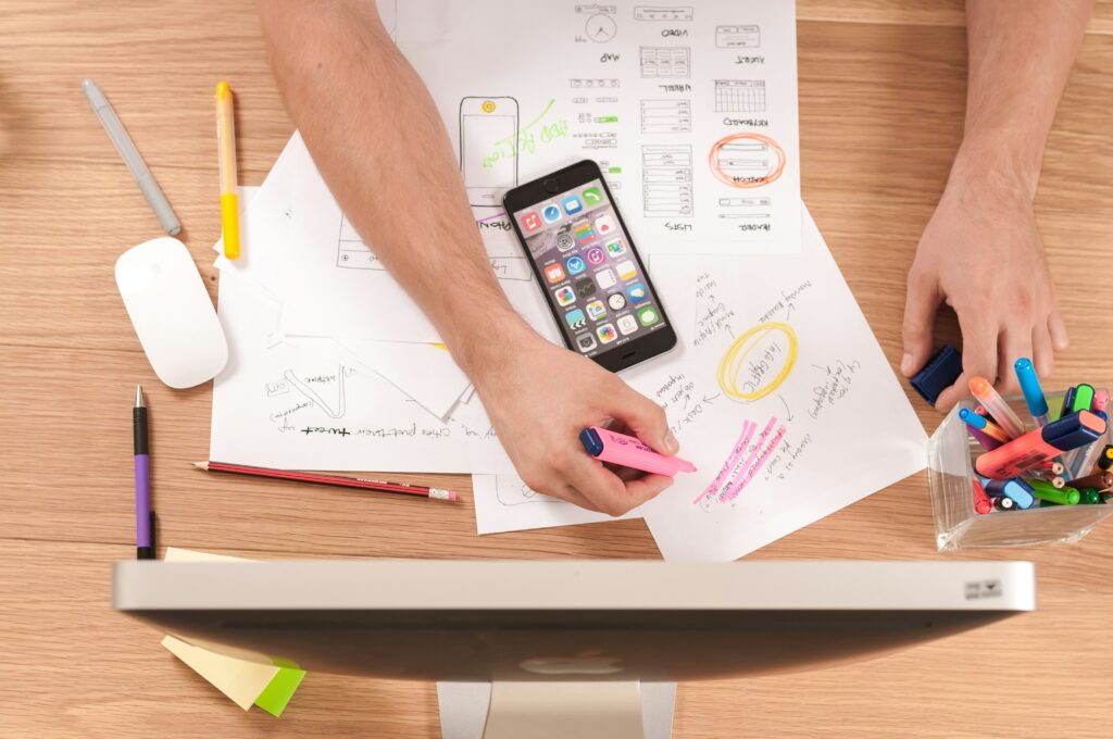Plan view of a desk with a PC, a mobile phone, stationery, several A4 sheets with hand-written notes and drawings, and two hands holding a highlighter pen - DevTeam.Space